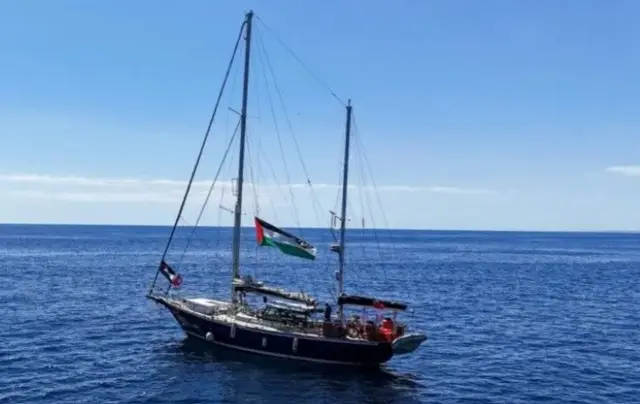 مشهد من طائرة بدون طيار لسفينة مادلين، سفينة المساعدات بإدارة تحالف أسطول الحرية. السفينة حملة العلم الفلسطيني. البحر أزرق صافٍ والسماء صافية نهاراً.